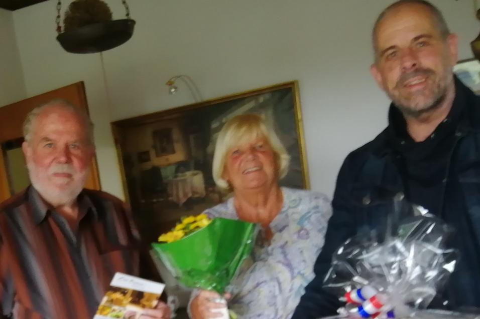 Drei Personen stehen in einem Raum, eine h&auml;lt einen Blumenstrau&szlig;, die anderen Geschenke und eine Brosch&uuml;re.