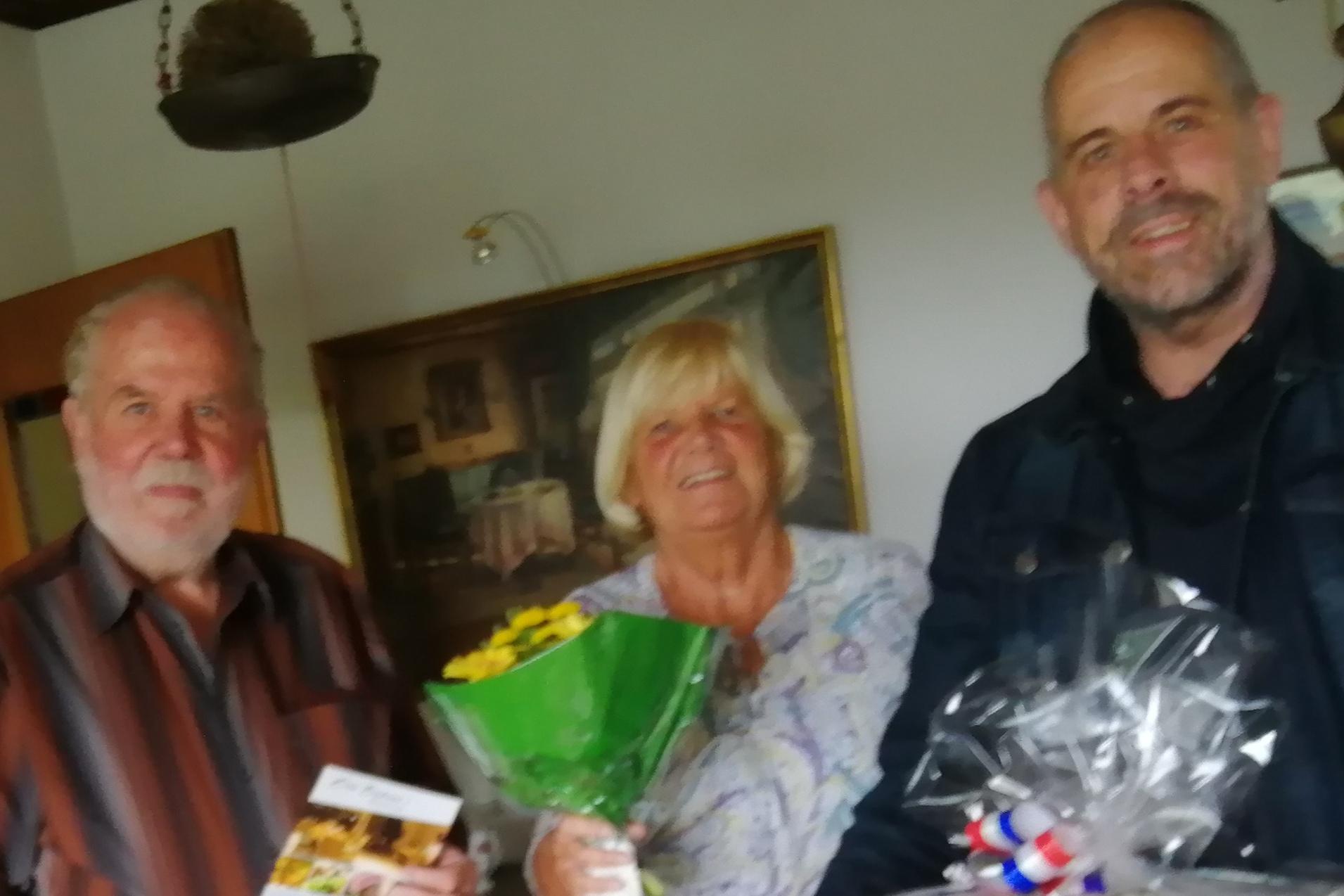 Drei Personen stehen in einem Raum, eine hält einen Blumenstrauß, die anderen Geschenke und eine Broschüre.