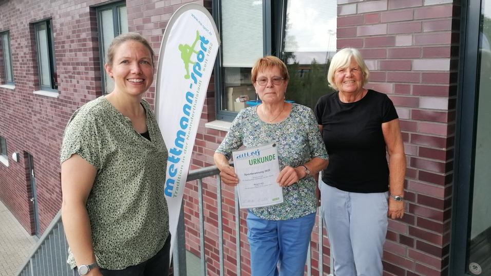 Drei Frauen stehen vor einem Backsteingebäude, eine hält eine Urkunde, im Hintergrund ein Werbebanner für "mettmann-sport".