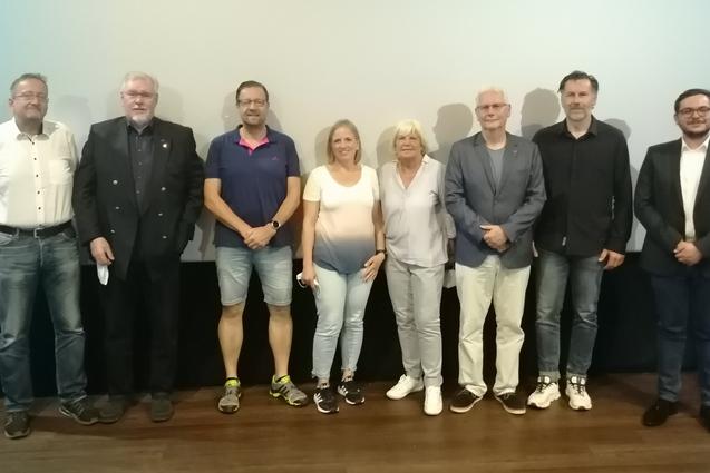 Gruppenaufnahme von acht Personen vor einem hellen Hintergrund in einem Veranstaltungsraum.