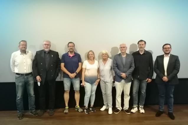 Gruppenaufnahme von acht Personen vor einem hellen Hintergrund in einem Veranstaltungsraum.