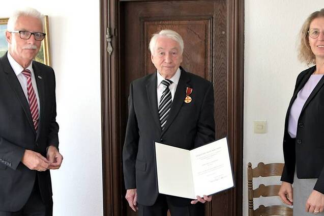 Drei Personen stehen in einem Raum; ein älterer Mann mit Medaille hält ein Zertifikat, umgeben von zwei anderen Erwachsenen.