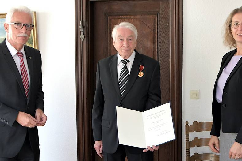 Drei Personen stehen in einem Raum; ein älterer Mann mit Medaille hält ein Zertifikat, umgeben von zwei anderen Erwachsenen.