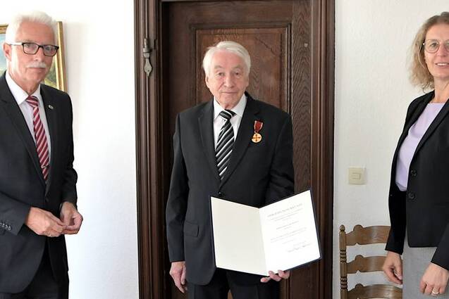 Drei Personen stehen in einem Raum; ein &auml;lterer Mann mit Medaille h&auml;lt ein Zertifikat, umgeben von zwei anderen Erwachsenen.