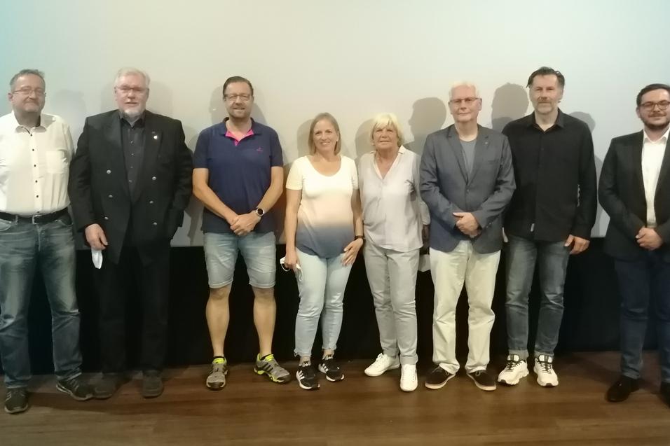 Gruppenaufnahme von acht Personen vor einem hellen Hintergrund in einem Veranstaltungsraum.