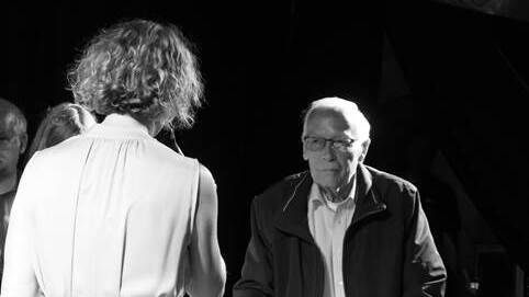 Ein älterer Mann mit Gehstützen steht einer Frau in einem weißen Kleid gegenüber. Schwarz-Weiß-Aufnahme.