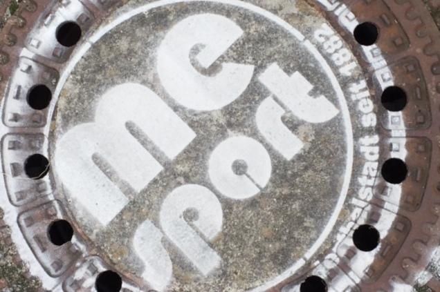 Gussdeckel mit der Aufschrift „me sport“ und mehreren Löchern, umgeben von Betonplatten.
