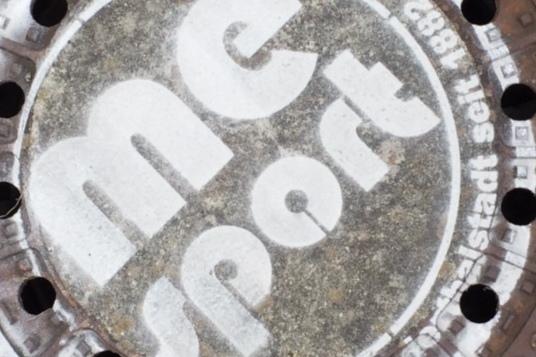 Gussdeckel mit der Aufschrift &bdquo;me sport&ldquo; und mehreren L&ouml;chern, umgeben von Betonplatten.
