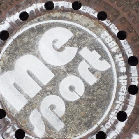 Gussdeckel mit der Aufschrift „me sport“ und mehreren Löchern, umgeben von Betonplatten.