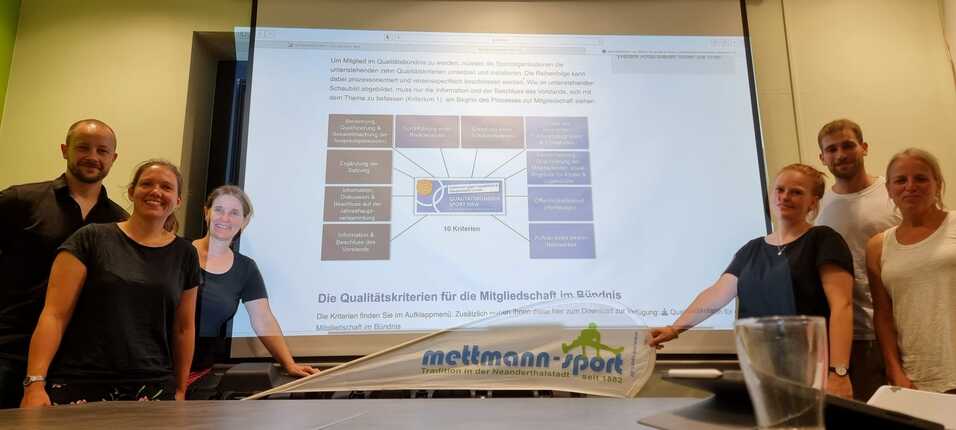 Sechs Personen stehen vor einem Präsentationsbildschirm mit Qualitätskriterien für den Sport NRW.