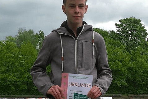 Junger Athlet auf dem Siegertreppchen, hält Urkunde in der Hand, mit Sportplatz im Hintergrund und bewölktem Himmel.