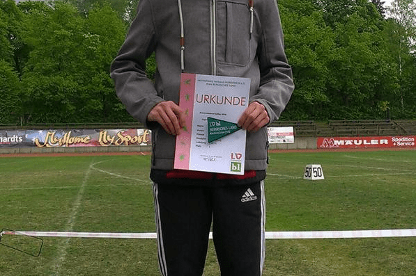 Junger Athlet auf dem Siegertreppchen, hält Urkunde in der Hand, mit Sportplatz im Hintergrund und bewölktem Himmel.