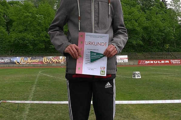 Junger Athlet auf dem Siegertreppchen, hält Urkunde in der Hand, mit Sportplatz im Hintergrund und bewölktem Himmel.