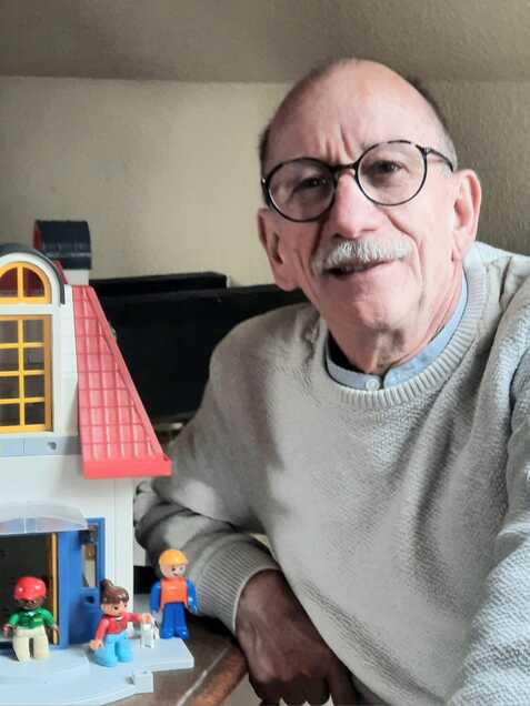 Älterer Mann sitzt neben einem bunten Spielhaus und mehreren Kunststofffiguren auf einem Tisch.