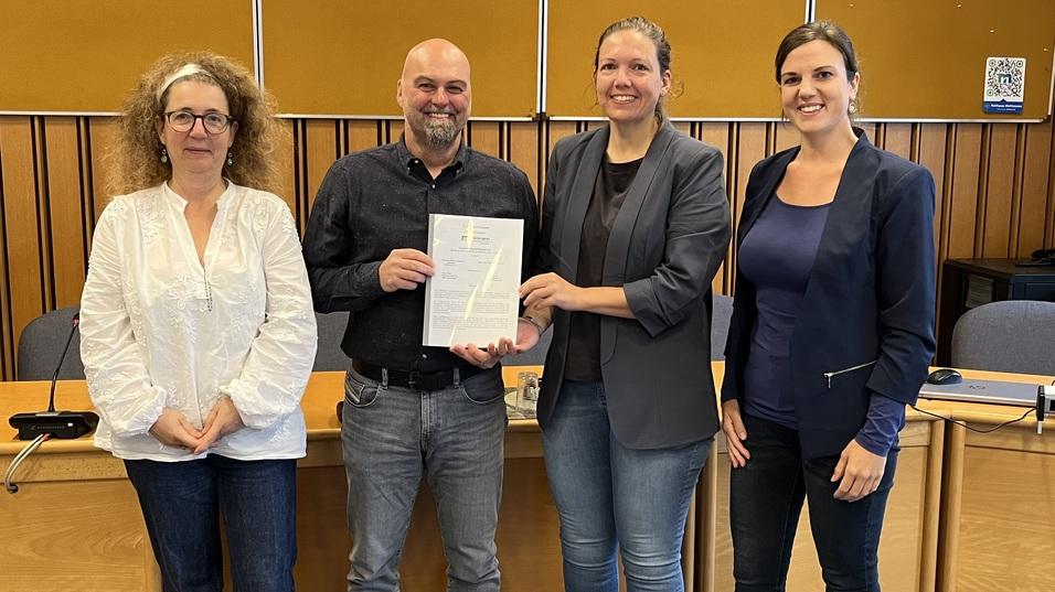 Vier Personen stehen in einem Konferenzraum, eine Person hält ein Dokument in der Hand, freundlich lächelnd.