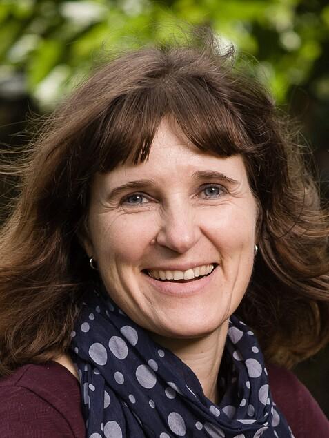 Lächelnde Frau mit langen, braunen Haaren und gepunktetem Schal vor unscharfem, grünem Hintergrund.