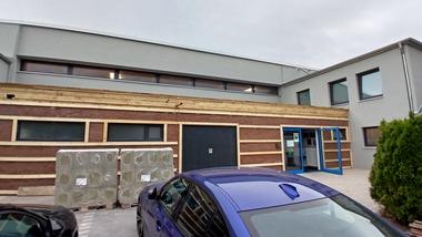 Eingang zu einem modernen Gebäude mit Holzverkleidung, zwei großen Fenstern und gelagerten Baustellenmaterialien.