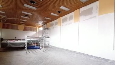 Unfertiger Raum mit Holzdecke, Gerüst und abgedecktem Tisch, Wände teilweise weiß und farblich grundiert.