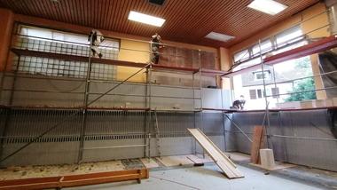 Arbeiter auf Gerüsten bei Renovierungsarbeiten in einem Raum mit großen Fenstern und Holzdecke.