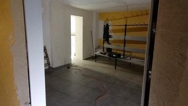 Blick durch einen offenen Türrahmen in einen lichtdurchfluteten Raum mit grauen Fliesen und gelben Wänden.