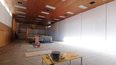 Großer, leerer Raum mit Holzboden, Baugerüst, Werkzeugen und abgedecktem Bodenbelag. Licht strömt von einem Fenster.