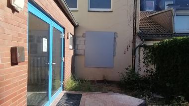 Eingangsbereich eines Gebäudes mit blauen Fenstern, unvollendeter Fliese, grauer Wand und verwildertem Garten.