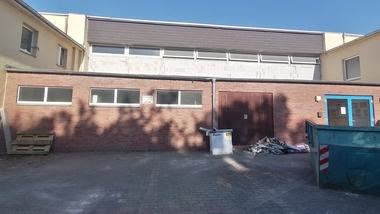 Rückansicht eines zweigeschossigen Gebäudes mit roten Ziegelwänden und Abfallcontainern auf einem gepflasterten Grundstück.