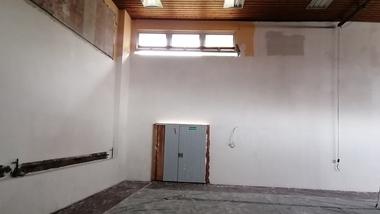 Leere Innenansicht eines Raumes mit grauer Tür, weißen Wänden und Holzdecke, auf dem Boden liegt eine Abdeckfolie.