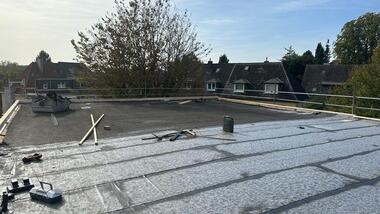 Blick auf eine flache Dachfläche mit Bauzubehör und einem Baum im Hintergrund. Wohnungen sind im Hintergrund sichtbar.