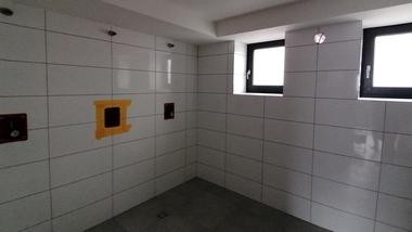 Blick in einen unfertigen Raum mit gefliesten Wänden, zwei Fenstern und Kabeln an der Decke. Gelbe Markierung für Steckdose.