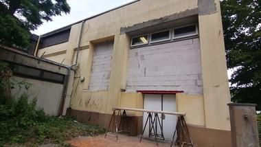 Verlassenes Gebäude mit unvollständigen Fenstern, Gerüst und verwildertem Hochbeet im Vordergrund.