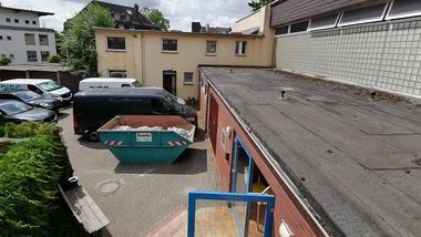 Blick auf einen Hinterhof mit verschiedenen Fahrzeugen, einem Container und einem flachen Gebäude unter bewölktem Himmel.