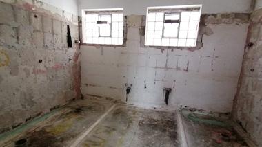 Leere, unrenovierte Raum mit zwei Fenstern, freiliegenden Leitungen und unvollständigem Estrichboden.