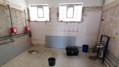 Unfertiger Raum mit freiliegenden Rohren, Fenstern, einem Eimer und einer schwarzen Schüssel auf dem Boden.