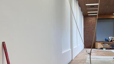 Blick auf eine weiße Wand in einem Raum mit Holzdecke und unvollendeten Bodenbelägen. Ein Gerüst steht bereit.