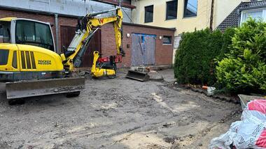 Baustelle mit gelbem Bagger und schwerem Verdichter auf unbefestigtem Untergrund, umgeben von Gebäuden und Grünflächen.