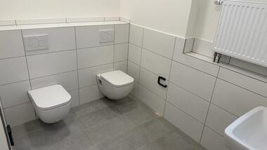 Modernes, helles, gefliestes WC mit zwei hängenden Toiletten und zwei Waschbecken an der Wand.