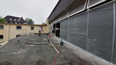 Arbeiter renovieren eine Dachterrasse mit Holzplanken und Werkzeug neben einer Gebäudefassade mit neuen Fenstern.