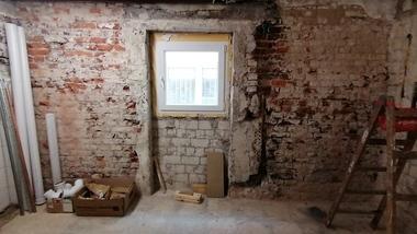 Rohbau eines Zimmers mit freiliegenden Ziegelwänden, Fenster und Baustellenmaterialien in der Ecke.