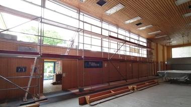 Blick in eine Sporthalle mit Gerüst, großen Fenstern und ungenutztem Equipment auf einem abgedeckten Boden.