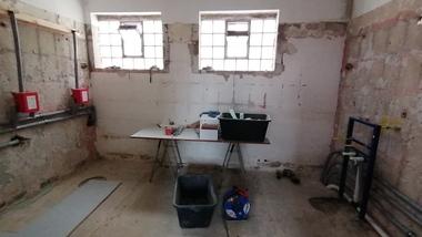 Blick in einen unrenovierten Raum mit Fenstern, einem Tisch, Werkzeugen und sichtbaren Rohren an der Wand.