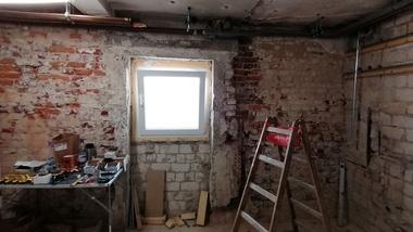 Rohbau eines Raums mit freiliegenden Ziegelwänden, Fenster, Leiter und Werkzeug auf einem Tisch.