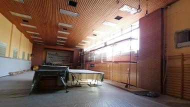 Renovierungsarbeiten in einer Sporthalle mit Gerüst, abgedeckten Tischen und Stoffbodenbelag. Licht fällt durch große Fenster.