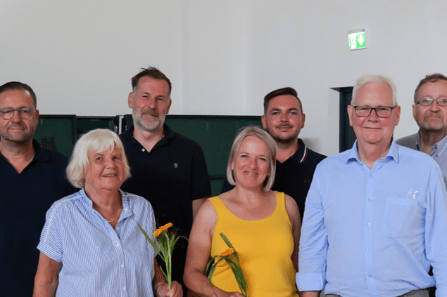 Gruppe von sieben Personen in einer Sporthalle, einige halten Blumen und Geschenke, beide Geschlechter vertreten.