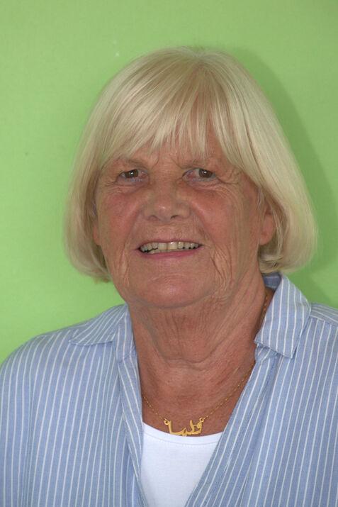 Porträt einer lächelnden älteren Frau mit blonden Haaren, im gestreiften Hemd vor grünem Hintergrund.