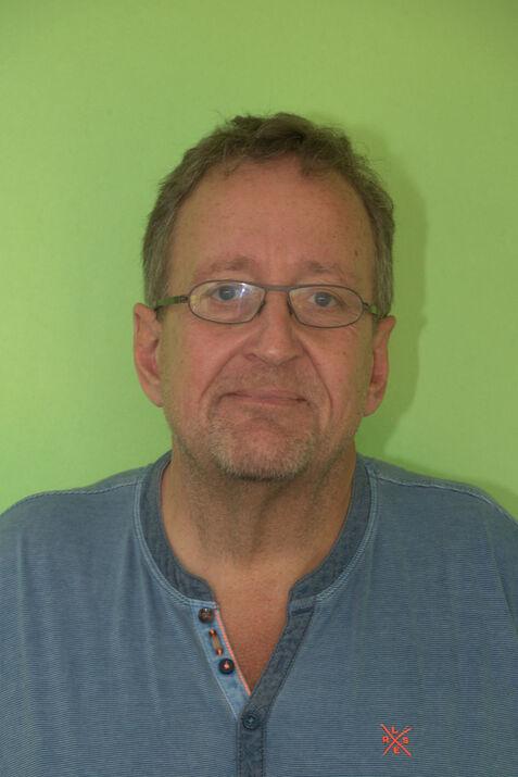 Portrait eines Mannes mit Brille und kurzen Haaren vor einer grünen Wand, wearing einem blauen Oberteil mit Knöpfen.