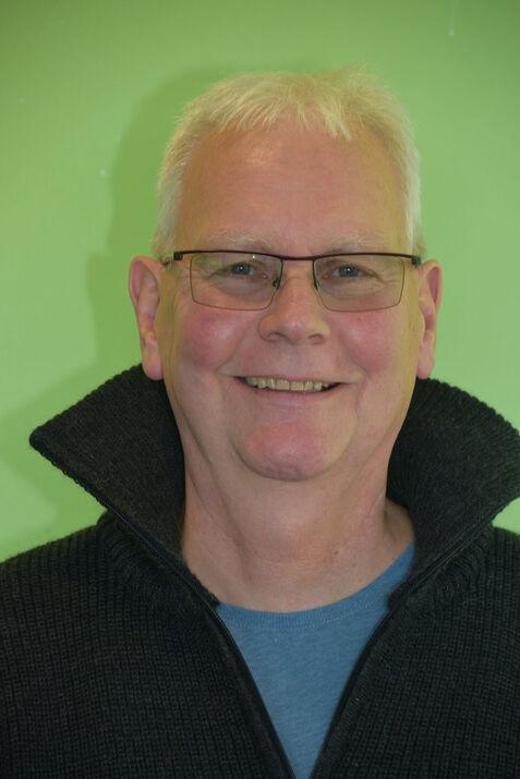 Porträt eines lächelnden Mannes mit Brille, grauen Haaren und schwarzem Pullover vor grünem Hintergrund.