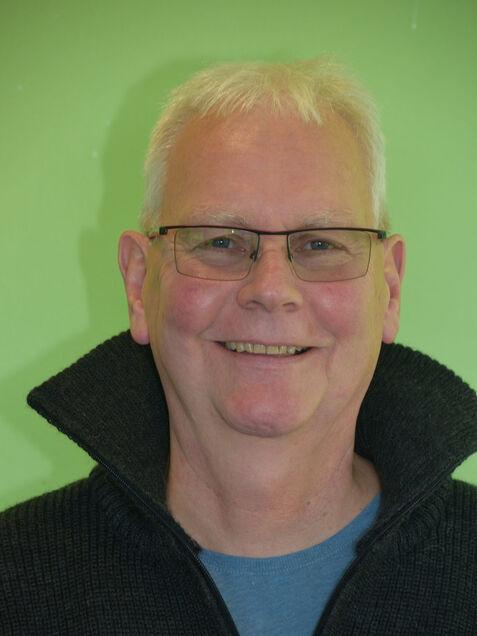 Porträt eines lächelnden Mannes mit Brille, grauen Haaren und schwarzem Pullover vor grünem Hintergrund.