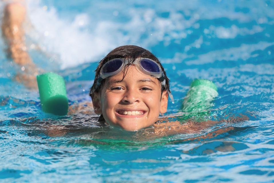 Lächelnder Junge mit Schwimmbrille schwimmt im Wasser, ausgestattet mit zwei grünen Schwimmhilfen.