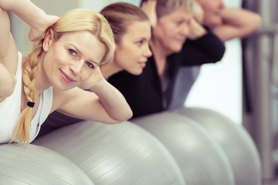 Eine Gruppe von fünf Personen übt auf Gymnastikbällen, einige halten die Hände hinter dem Kopf.